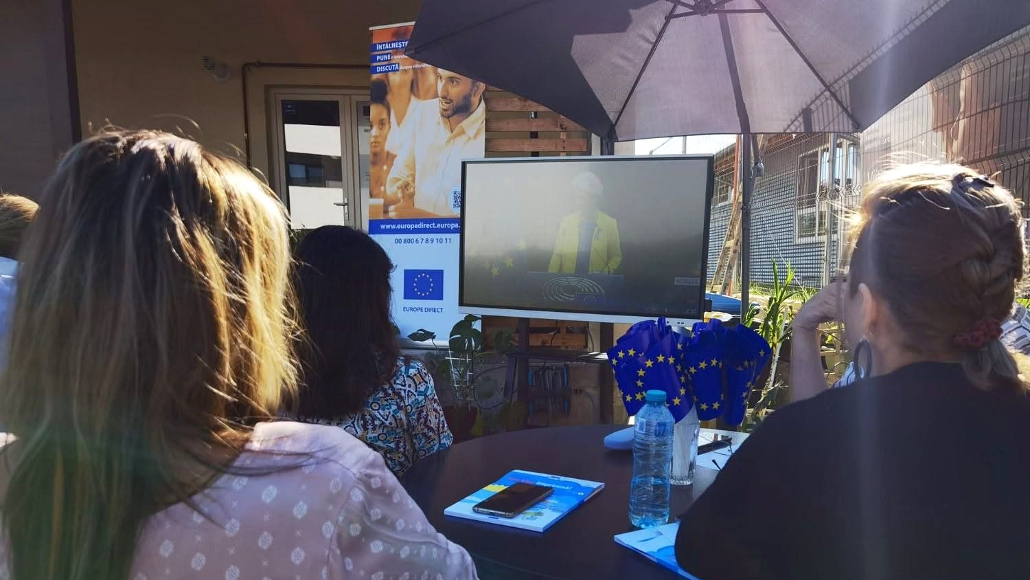 Participanți așezați la o masă în curtea centrului urmăresc pe un ecran discursul privind starea Uniunii; pe masă sunt stegulețe ale Uniunii Europene.